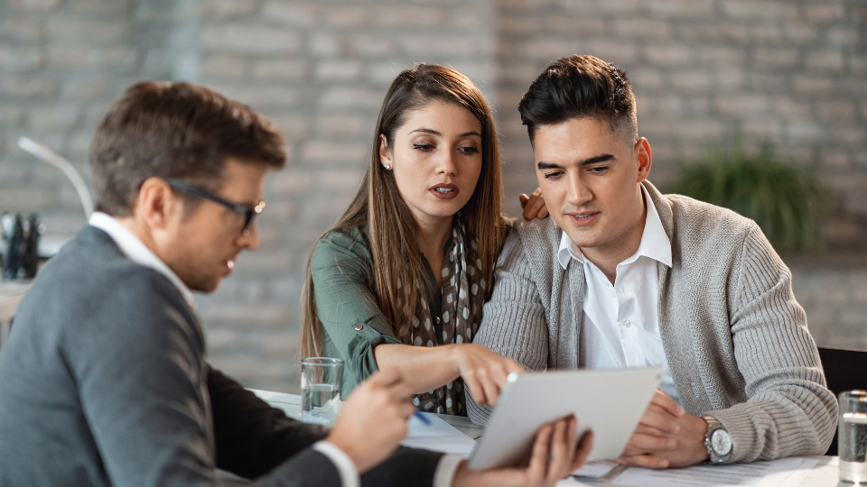 A lender reviews two borrowers’ financial documents across a desk to verify assets for mortgage eligibility
