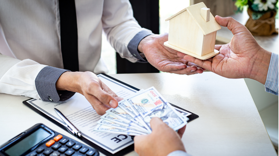 A person hands over cash and receives a wooden house model on a desk with documents and a calculator