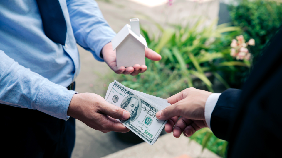 A person hands over cash while another person holds a small model house outdoors