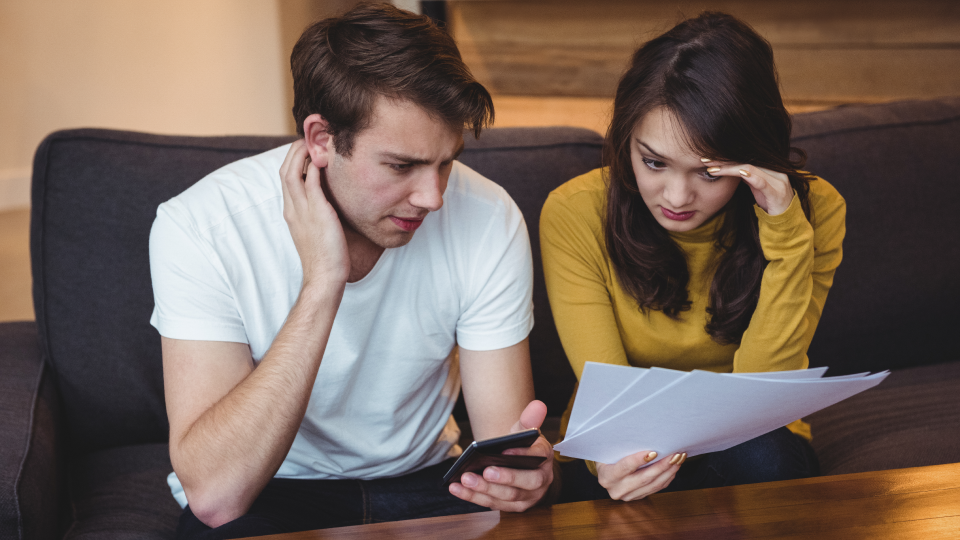 A couple sits on a couch, looking concerned while reviewing documents and a smartphone
