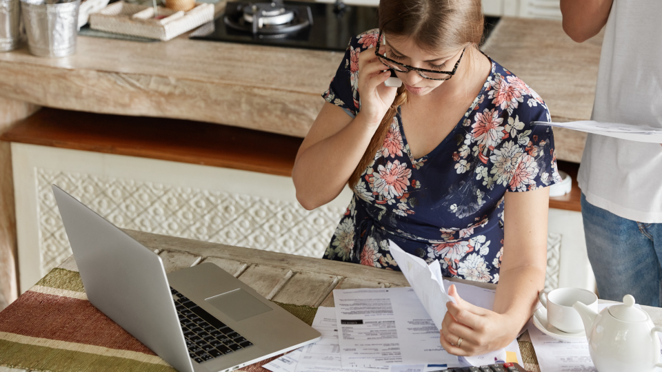A woman talks on the phone while reviewing documents at a kitchen table with a laptop and paperwork