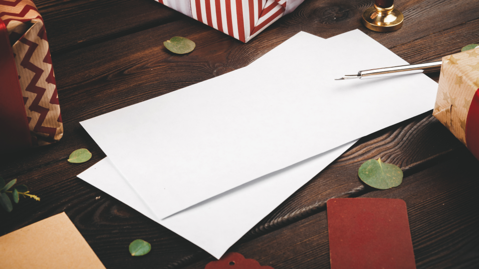 A blank envelope and pen sit on a wooden table next to gifts and decorative leaves
