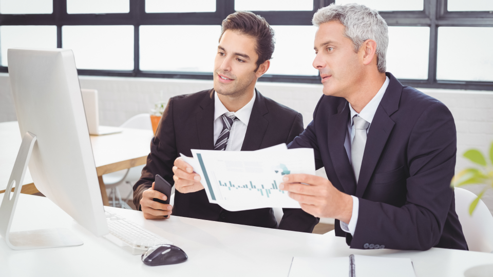 Two men in business attire review a printed financial report at a computer desk.