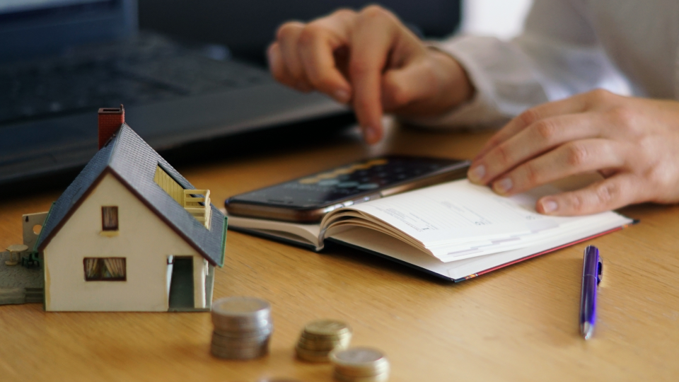 A person uses a calculator and smartphone next to a miniature house, notebook, and coins on a desk