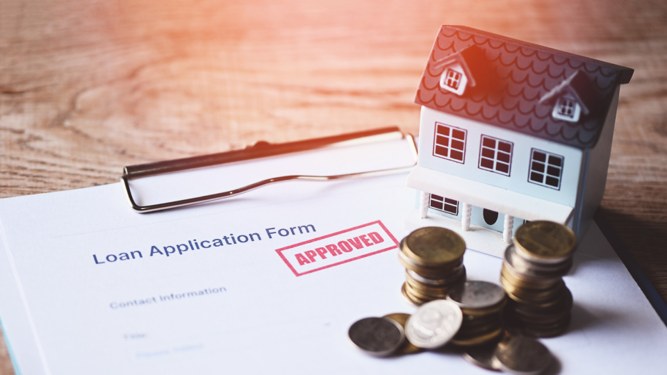 A loan application form with an approved stamp sits on a desk next to stacked coins and a small house model