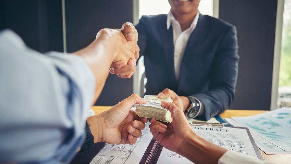 Two people hand a large amount of cash across a desk