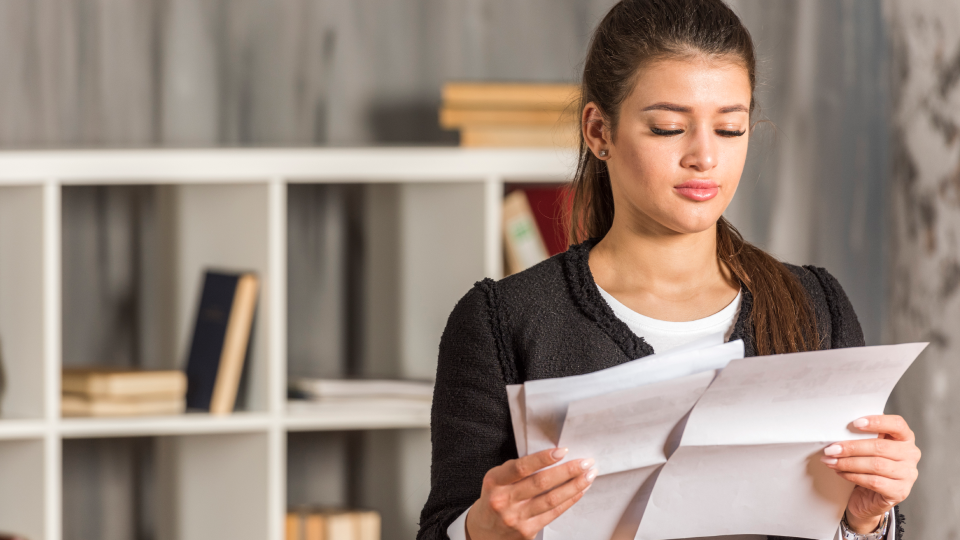 A woman stands in front of a bookshelf and reads several printed documents