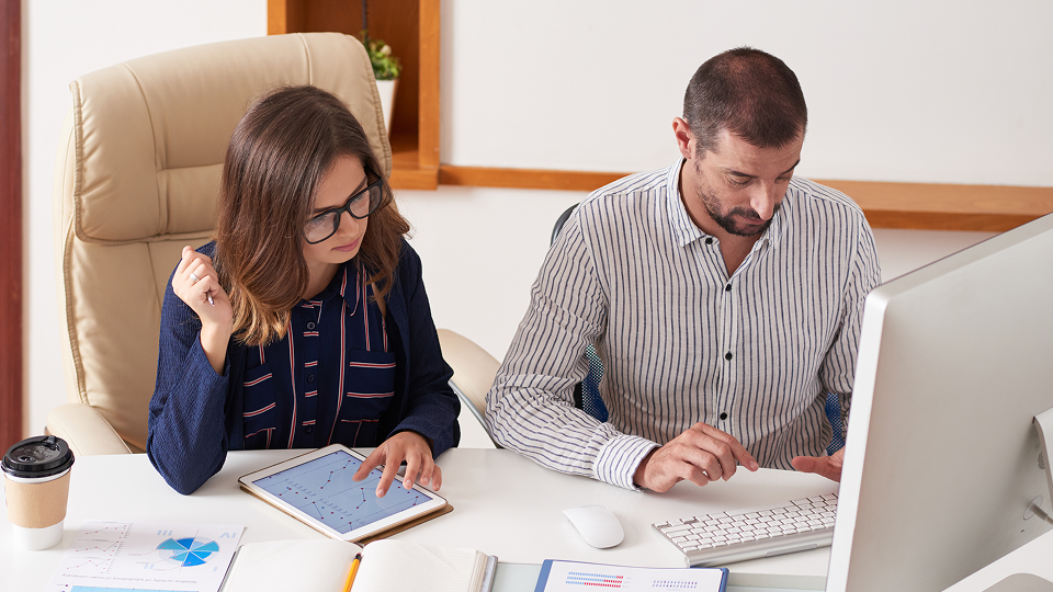 Two professionals review data on a computer and tablet at an office desk with charts and reports